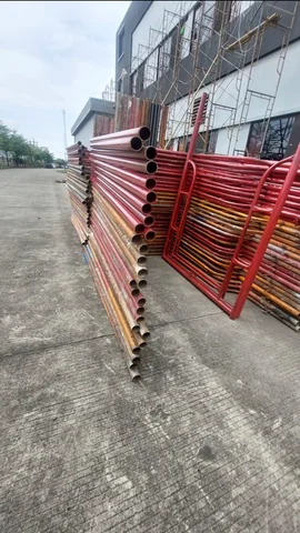 Tumpukan frame dan pipa scaffolding bercat merah bersandar di area bangunan